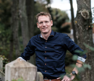 Portret van een ondernemer buiten in een groene omgeving, staand bij een hek, met bomen op de achtergrond.